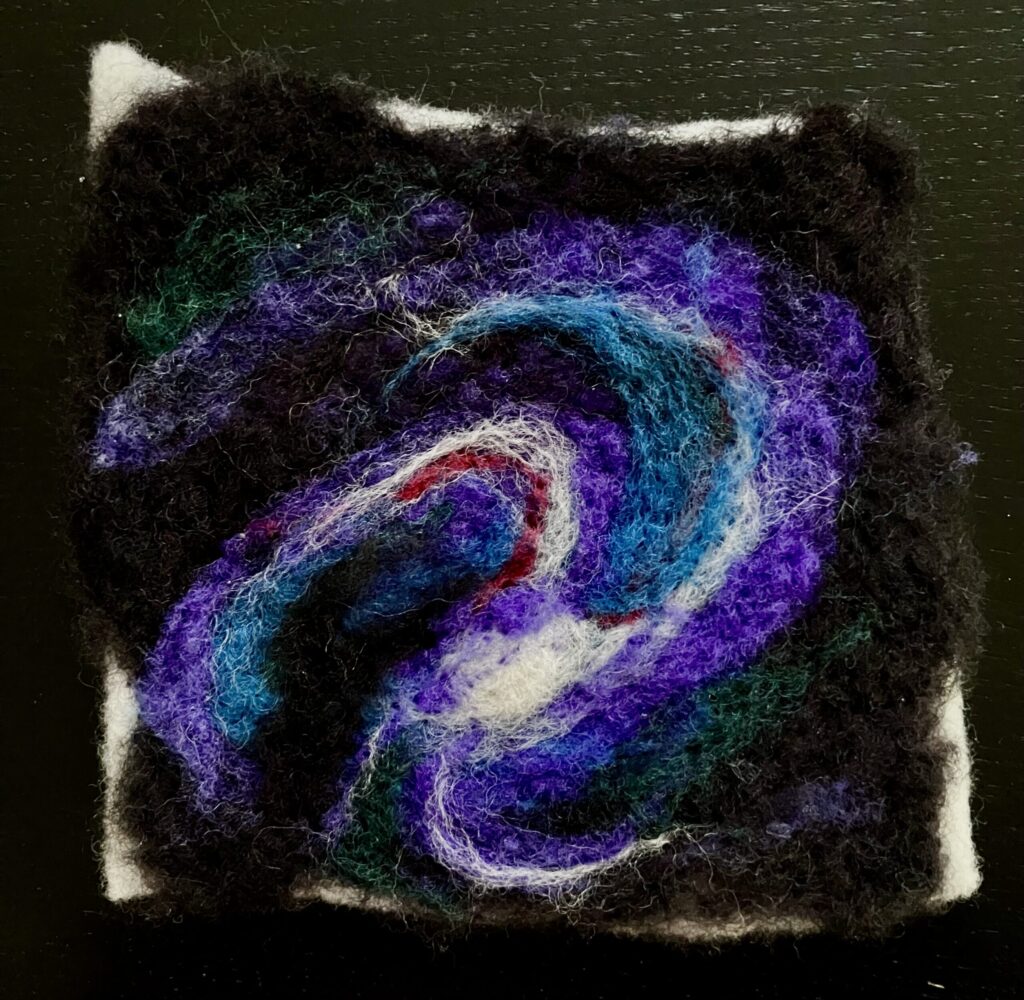 A needlefelted galaxy in black, white, purple, blue, and red.