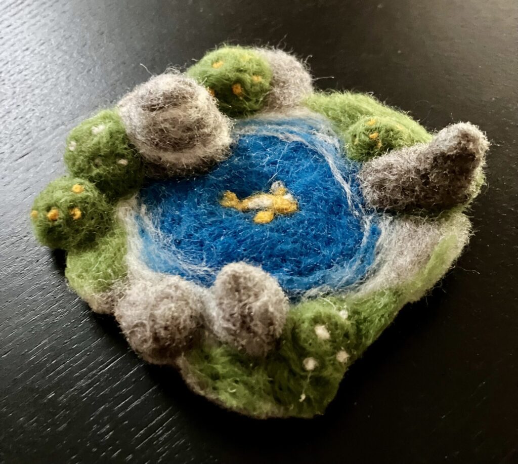 A needlefelted pool surrounded by stones and greenery, with a koi fish.