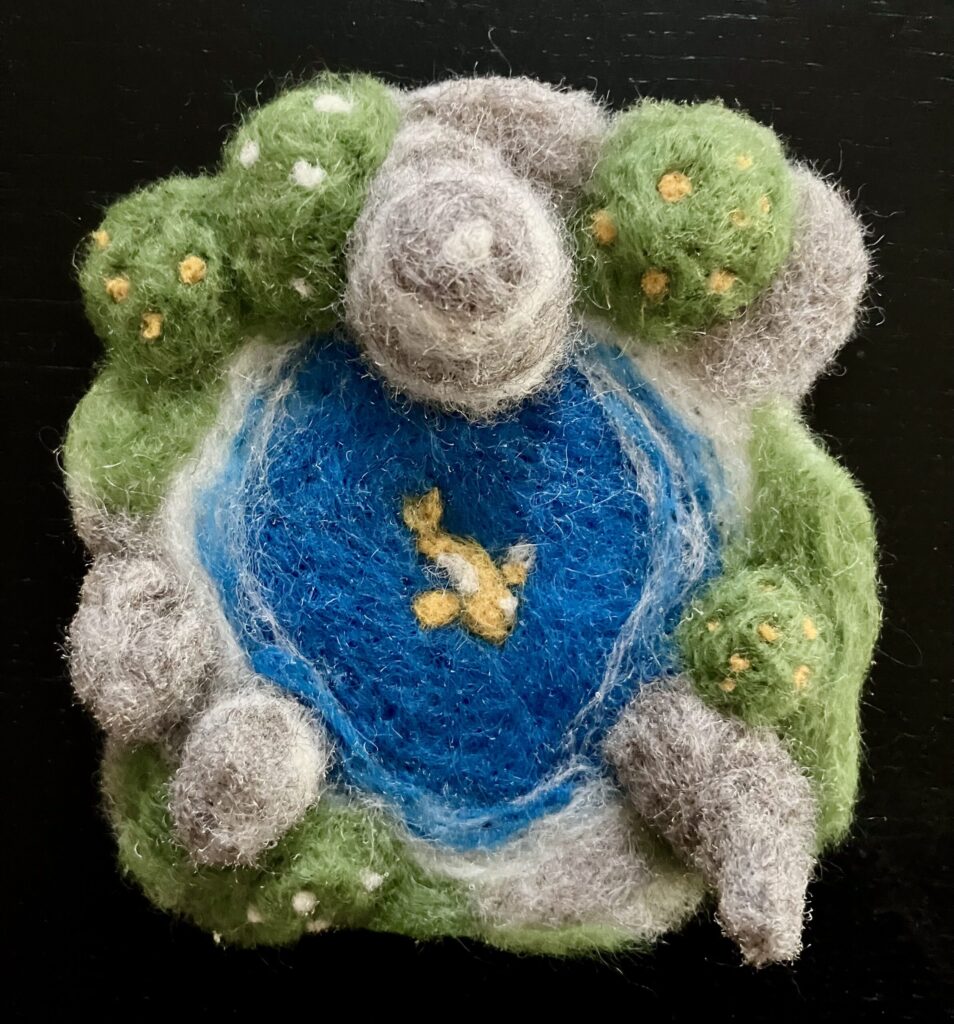 A needlefelted pool surrounded by stones and greenery, with a koi fish.