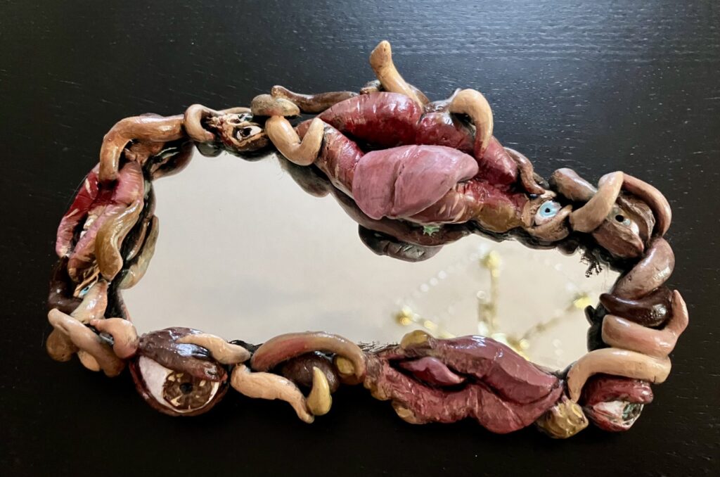 A mirror with a border of sculpted mouths, eyes, and fleshy tendrils.