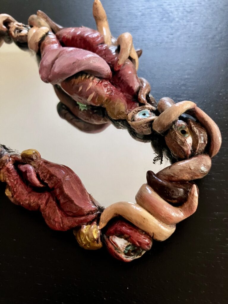 A mirror with a border of sculpted mouths, eyes, and fleshy tendrils.