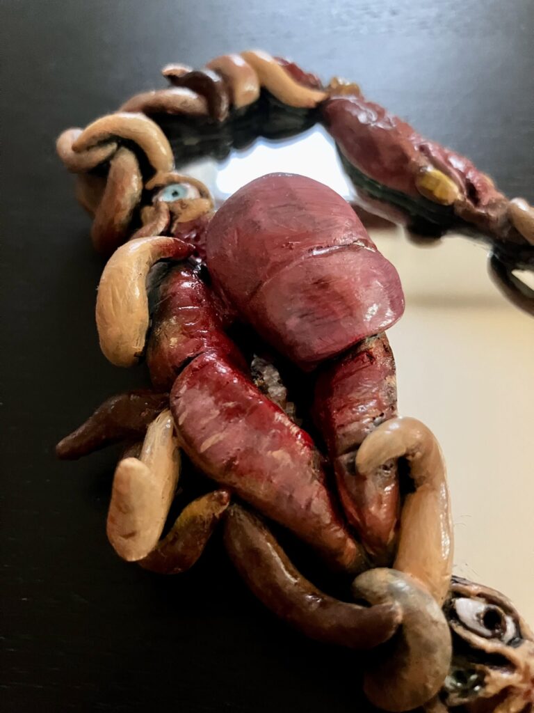 A mirror with a border of sculpted mouths, eyes, and fleshy tendrils.