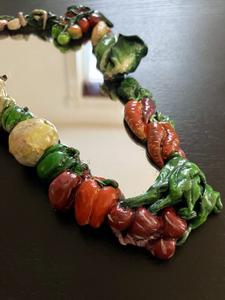A close up of a mirror bordered by painted, sculpted vegetables.