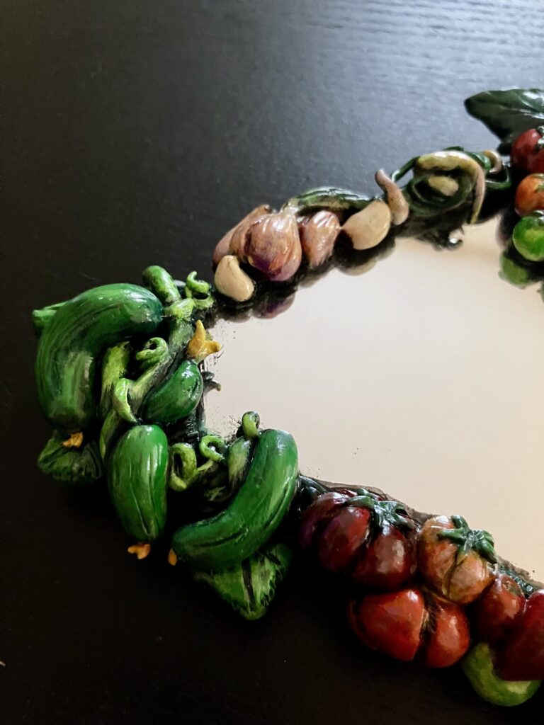 A close up of a mirror bordered by painted, sculpted vegetables.