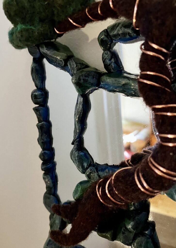 A photo of an assemblage of mirrors with sculpted faux stone borders and a needlefelted bonsai tree wired to them.