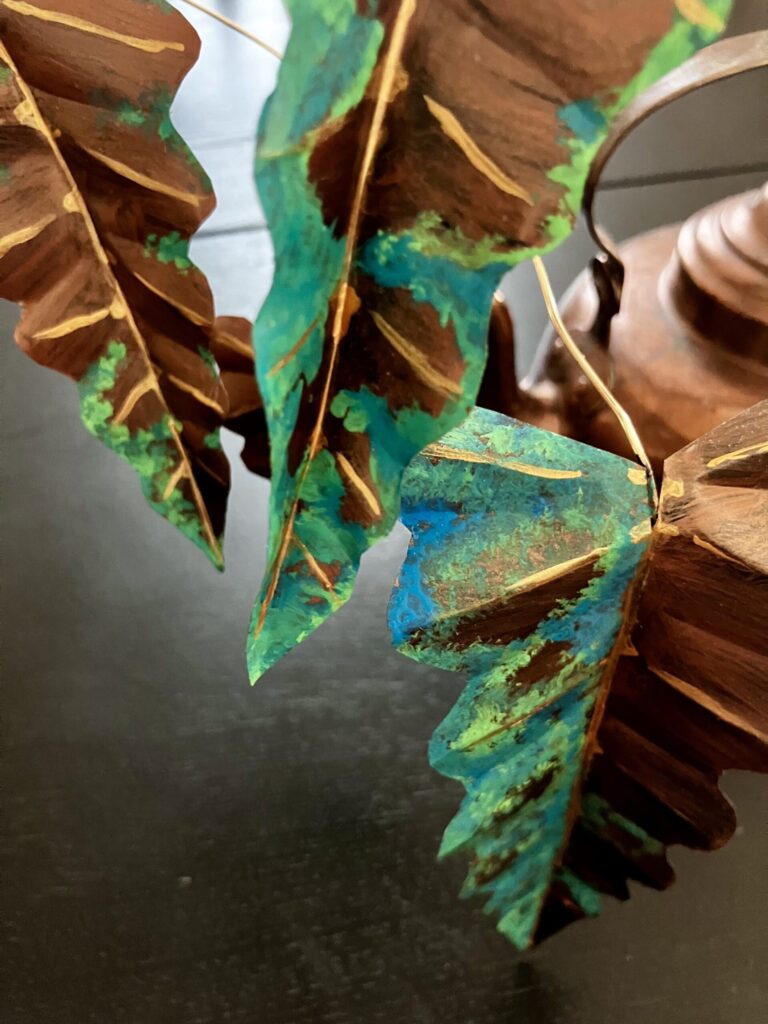 A photo of a sculpted copper colored alocasia growing out of a copper kettle.