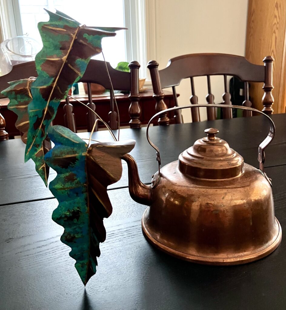 A photo of a sculpted copper colored alocasia growing out of a copper kettle.