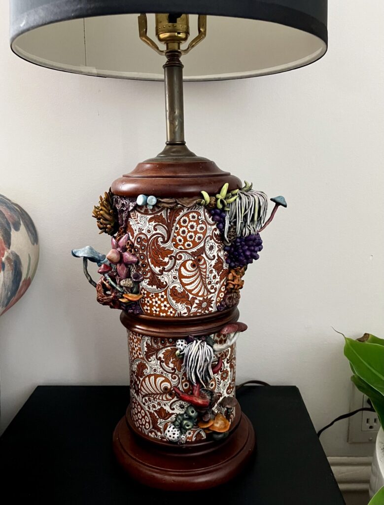 A photo of a tabletop lamp made of wood and ceramic with a dark orange paisley pattern. Sculpted mushrooms of a variety of kinds climb up the sides.
